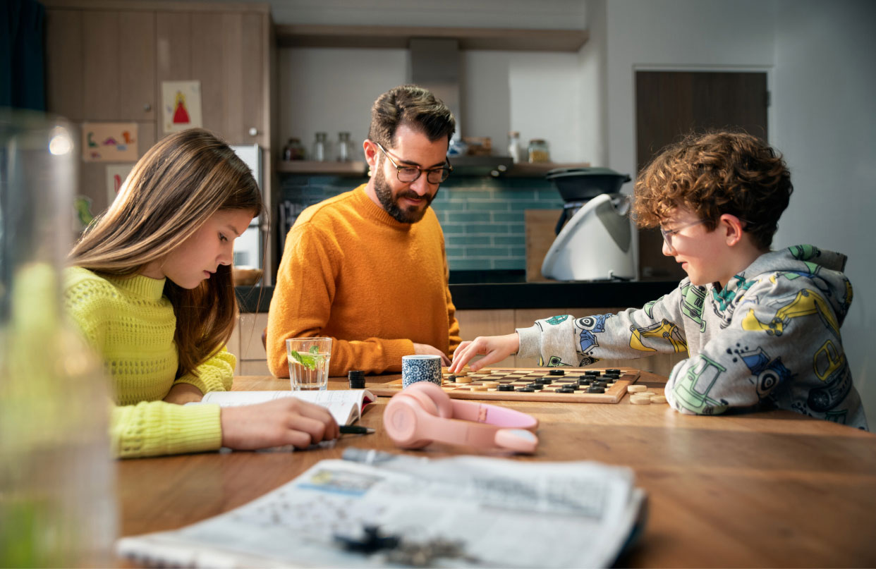 Father and children playing checkers and studying at a wooden kitchen table.